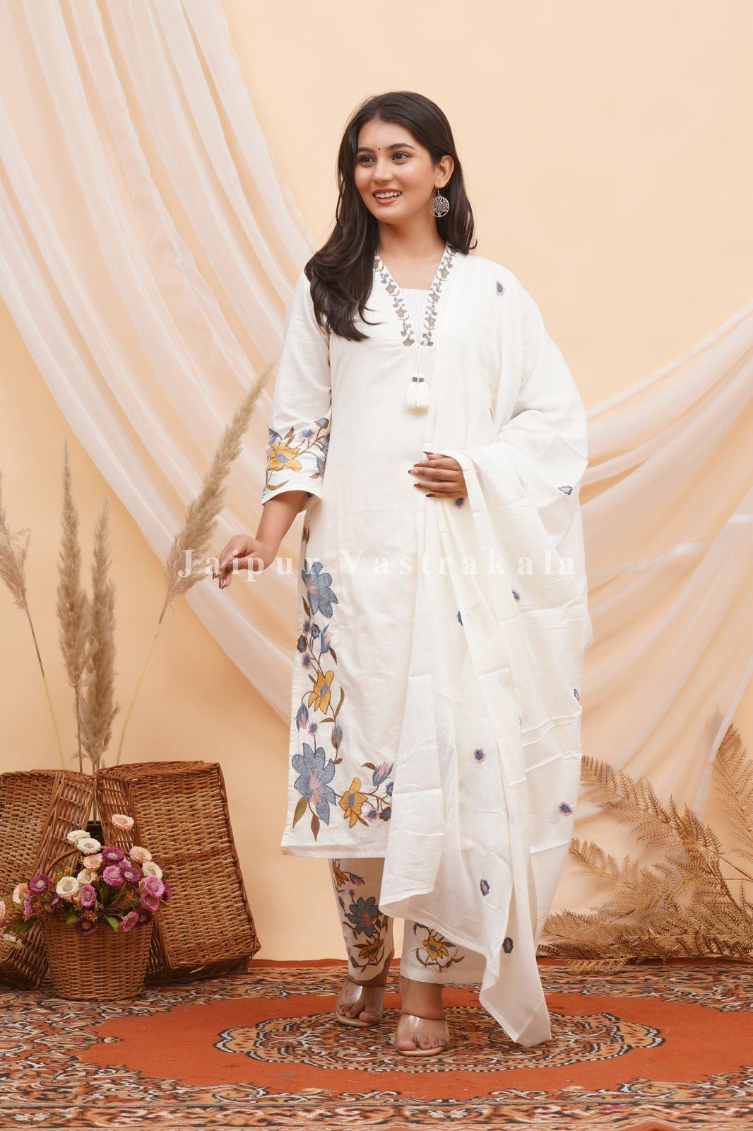 Woman in a white traditional outfit with floral patterns standing against a beige curtain.