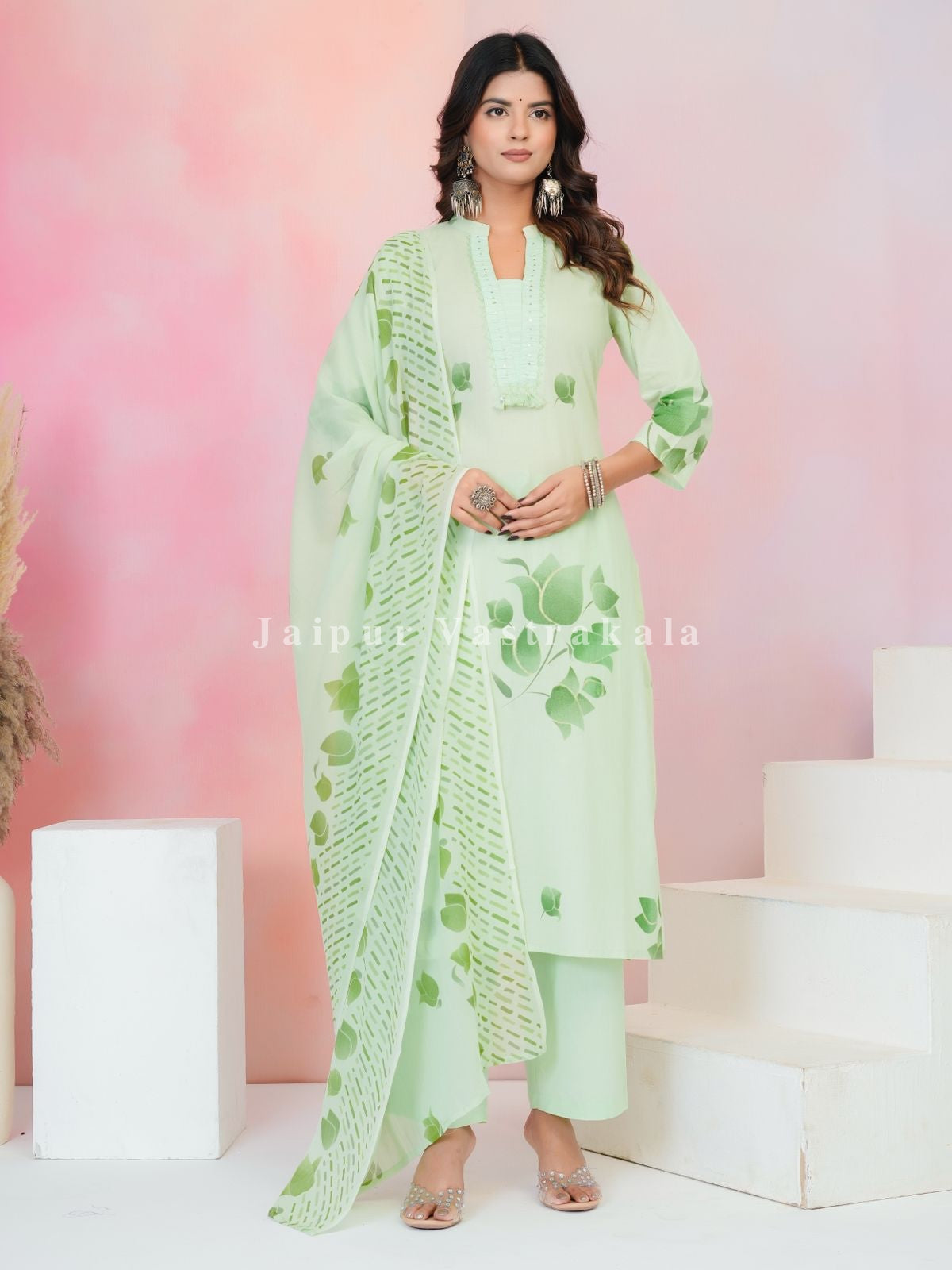 Woman wearing a light green traditional outfit with a floral pattern against a pink and white background.